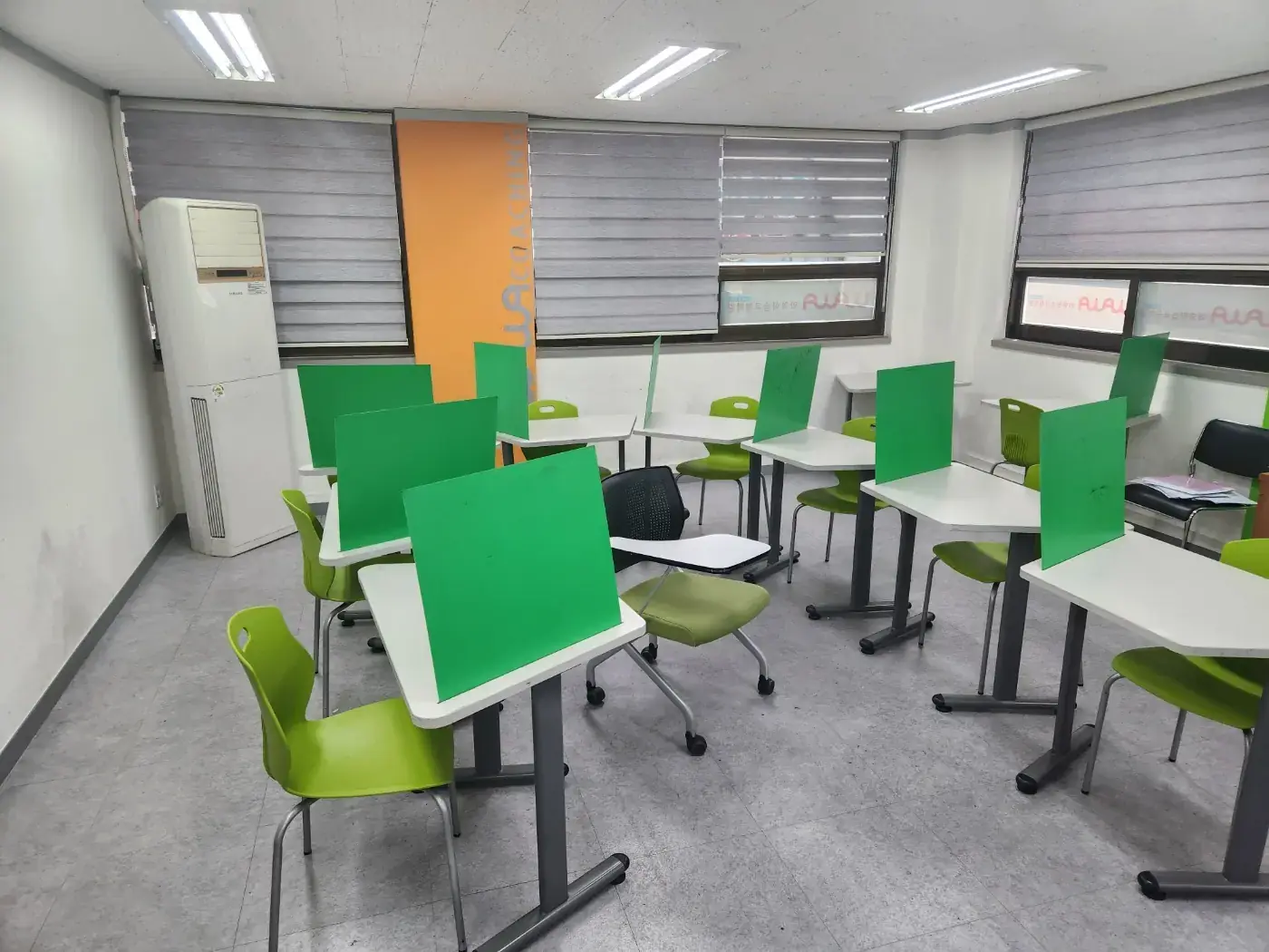 회색 블라인드 창과 주황색 와와 코칭 기둥 앞에 초록 칸막이 책상이 놓인 철산점 교실